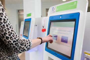 Touch screen with a womans hand pointing to it. On the screen is check in information.