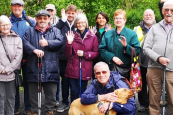 a group of people stand outside and wave at the camera. a dog is at the front of the group