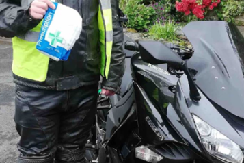 A man in a high visibility jacket stands next to a motorbike while holding a bag of prescription medication.