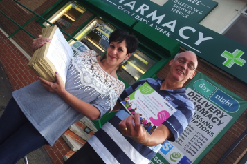 A man and a woman stand infront of a pharmacy. The woman holds a stack of questionnaires and the man holds a Healthwatch Dudley postcard.