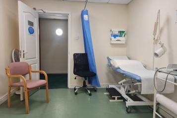 Photograph of a treatment room. Inside is a blue treatment couch, two chairs and medical equipment.